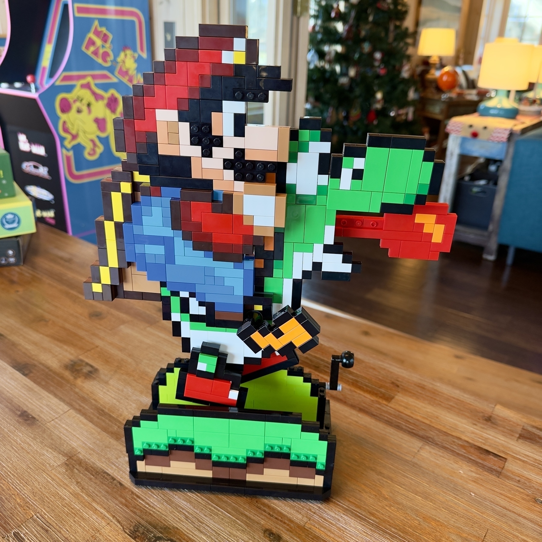 Auto-generated description: A block-style character resembling a plumber riding a green dinosaur, assembled from toy bricks, stands on a wooden surface with arcade machines and a Christmas tree in the background.