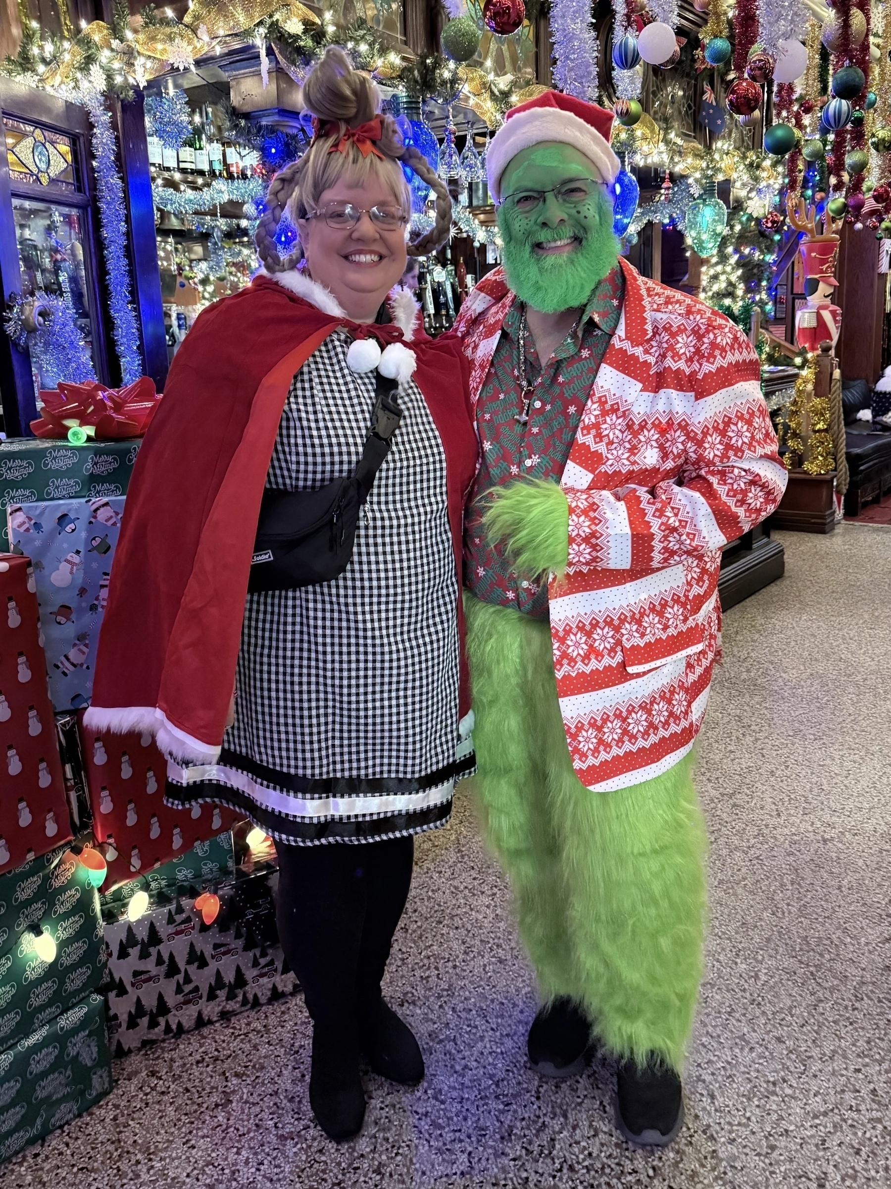 Auto-generated description: Two people in holiday-themed costumes, one dressed as a version of the Grinch and the other with festive attire, stand in a decorated room with presents and lights.