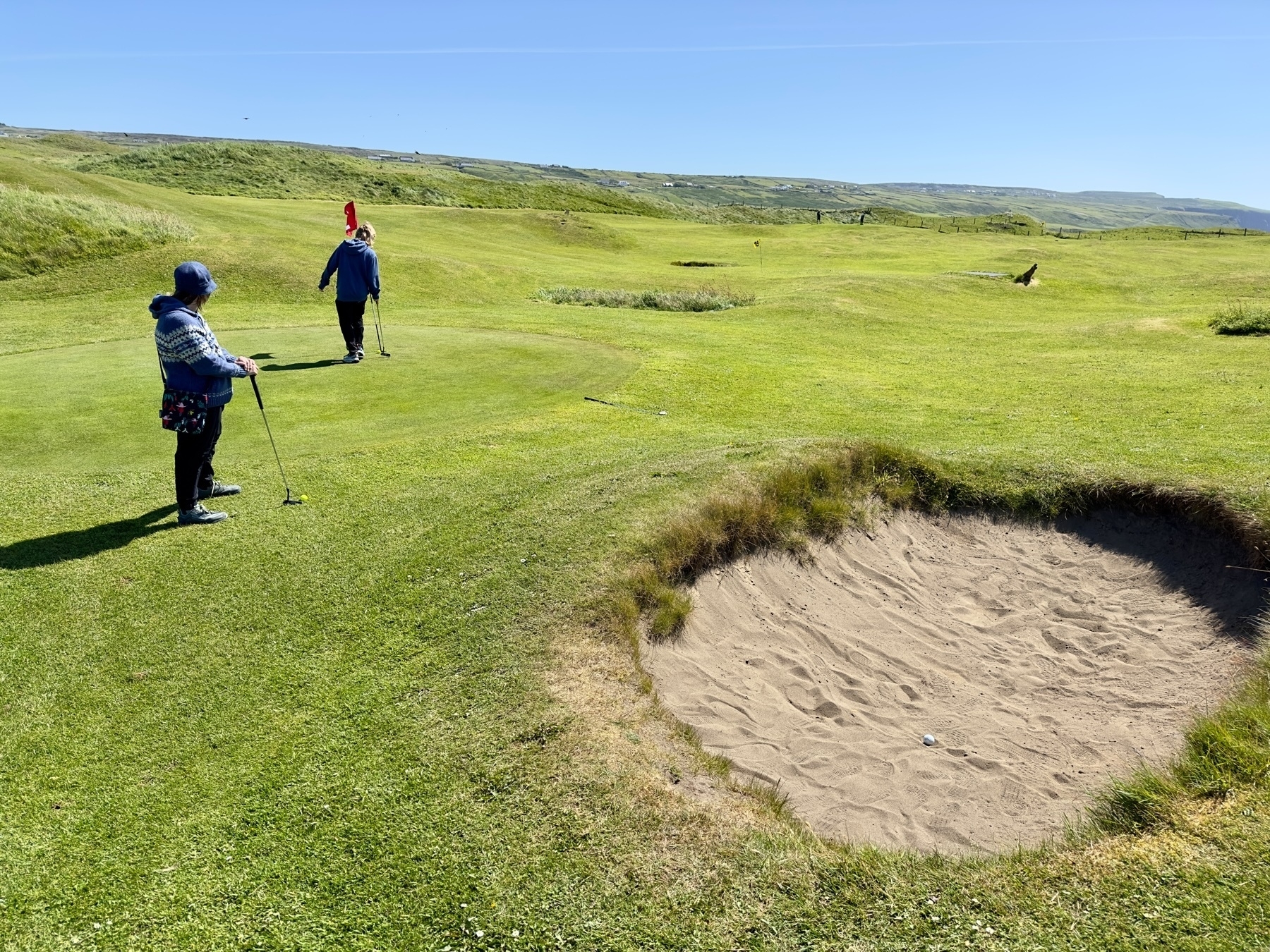 Auto-generated description: Two people are playing golf on a sunny day, with one preparing to putt near a sand bunker and the other standing near the flag on an open, grassy course.
