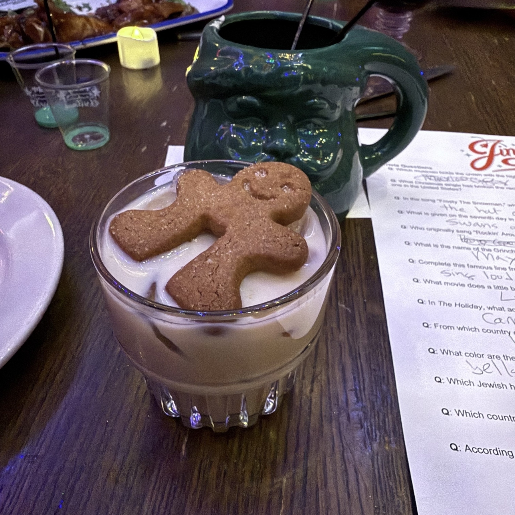 Auto-generated description: A gingerbread cookie is placed on the rim of a glass filled with a creamy beverage, set on a wooden table with a green ceramic mug nearby.