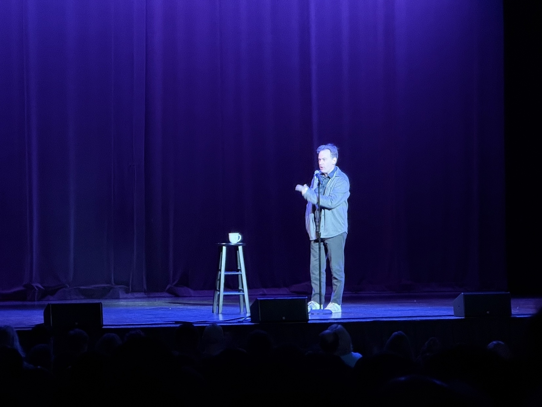 Auto-generated description: A comedian performs on a stage with a spotlight, standing near a stool with a cup on it.