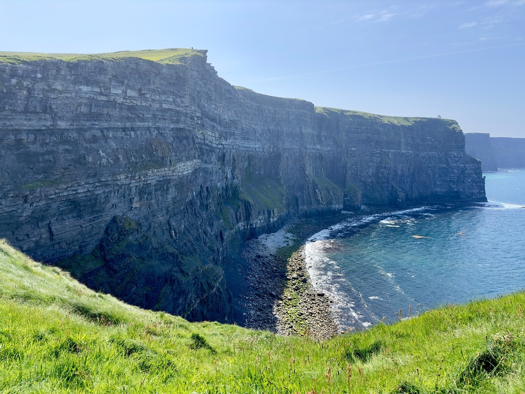 Auto-generated description: A majestic coastal cliff with layered rock formations overlooks a serene shoreline and vibrant blue sea under a clear sky.