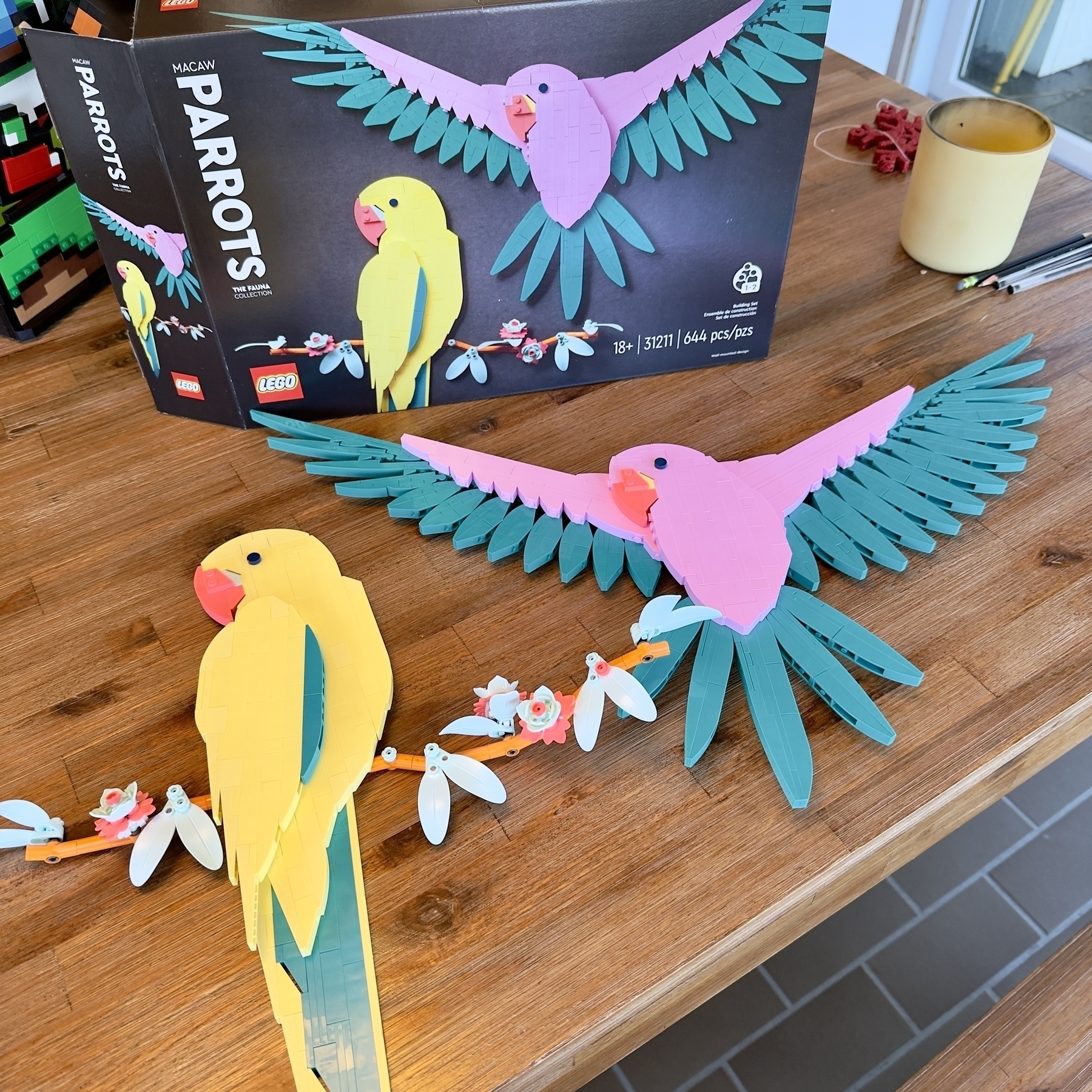 Auto-generated description: Two colorful LEGO macaws, one perched and another flying, are showcased on a wooden table alongside the building set box.