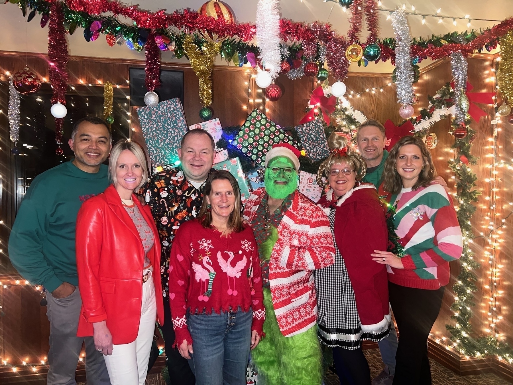 Auto-generated description: A group of people, wearing festive holiday sweaters, pose in front of a decorated Christmas-themed backdrop, with one person dressed as the Grinch.