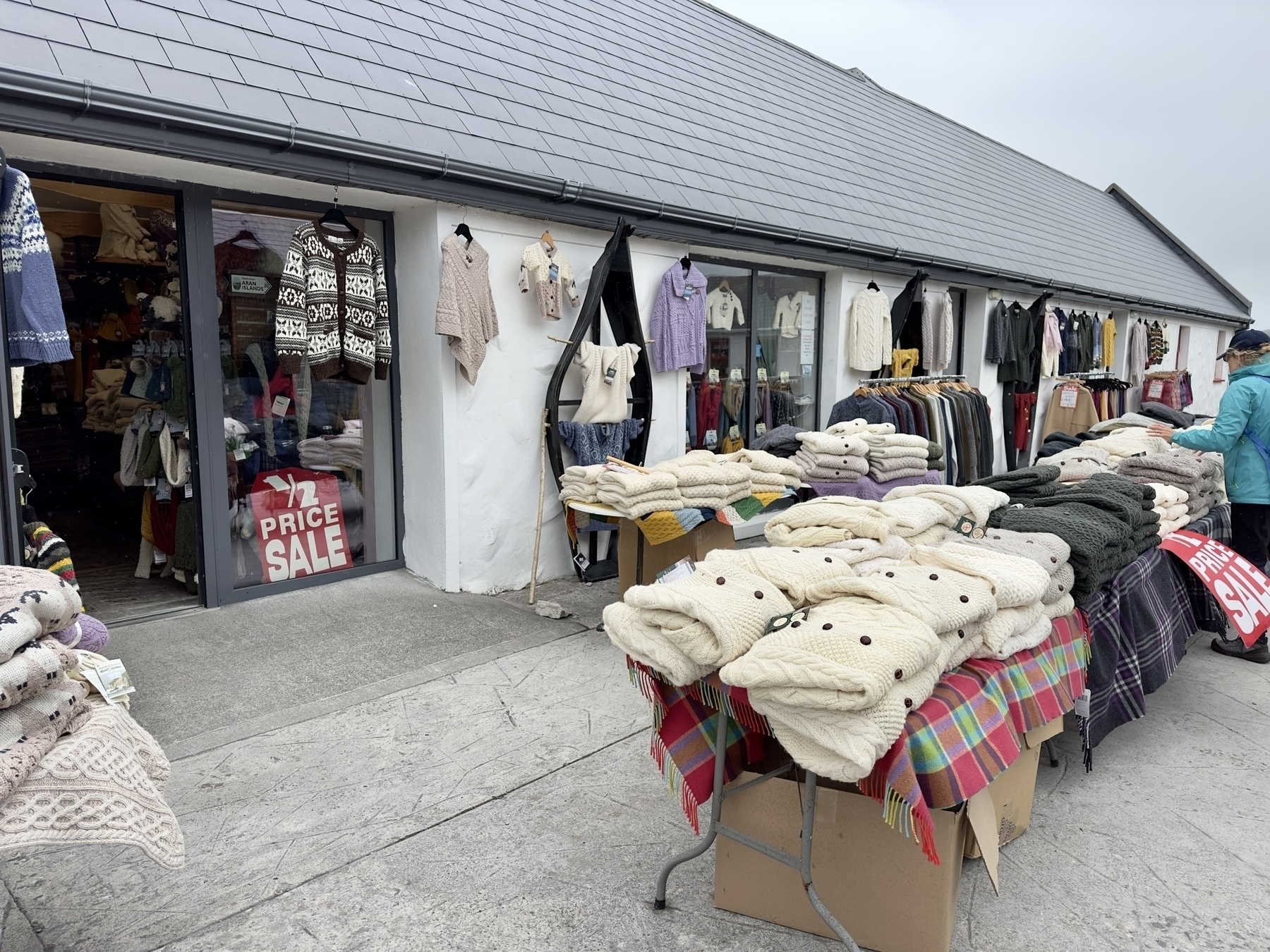 Auto-generated description: A market stall is displaying various sweaters and garments on tables, with additional clothing hanging on the walls and racks inside a building, and a Price Sale sign visible.