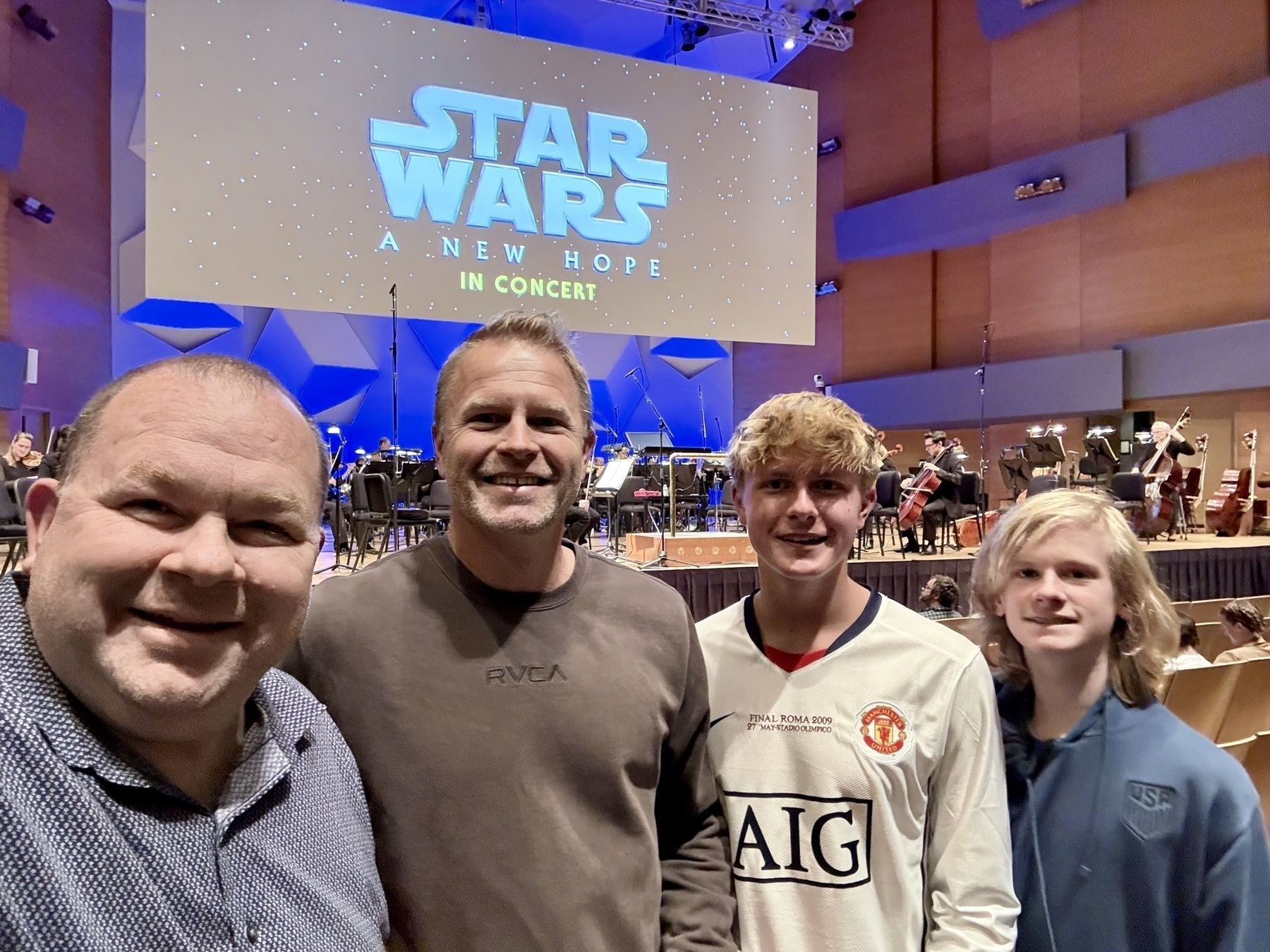 Auto-generated description: Four people are smiling and posing in front of a stage set for a Star Wars: A New Hope concert.