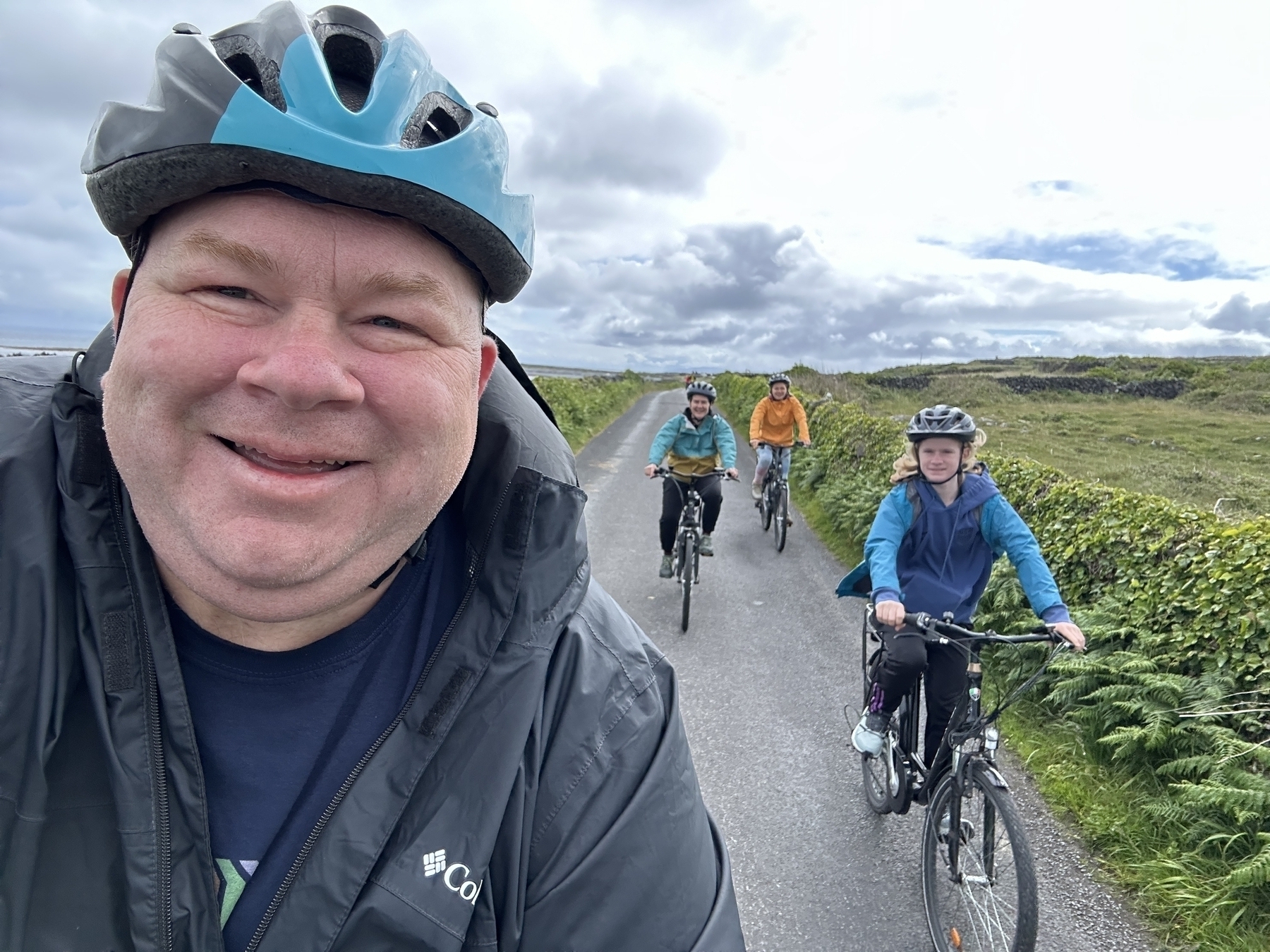 Auto-generated description: A group of people wearing helmets are riding bicycles on a narrow road with greenery on both sides, with one person taking a selfie from the front.