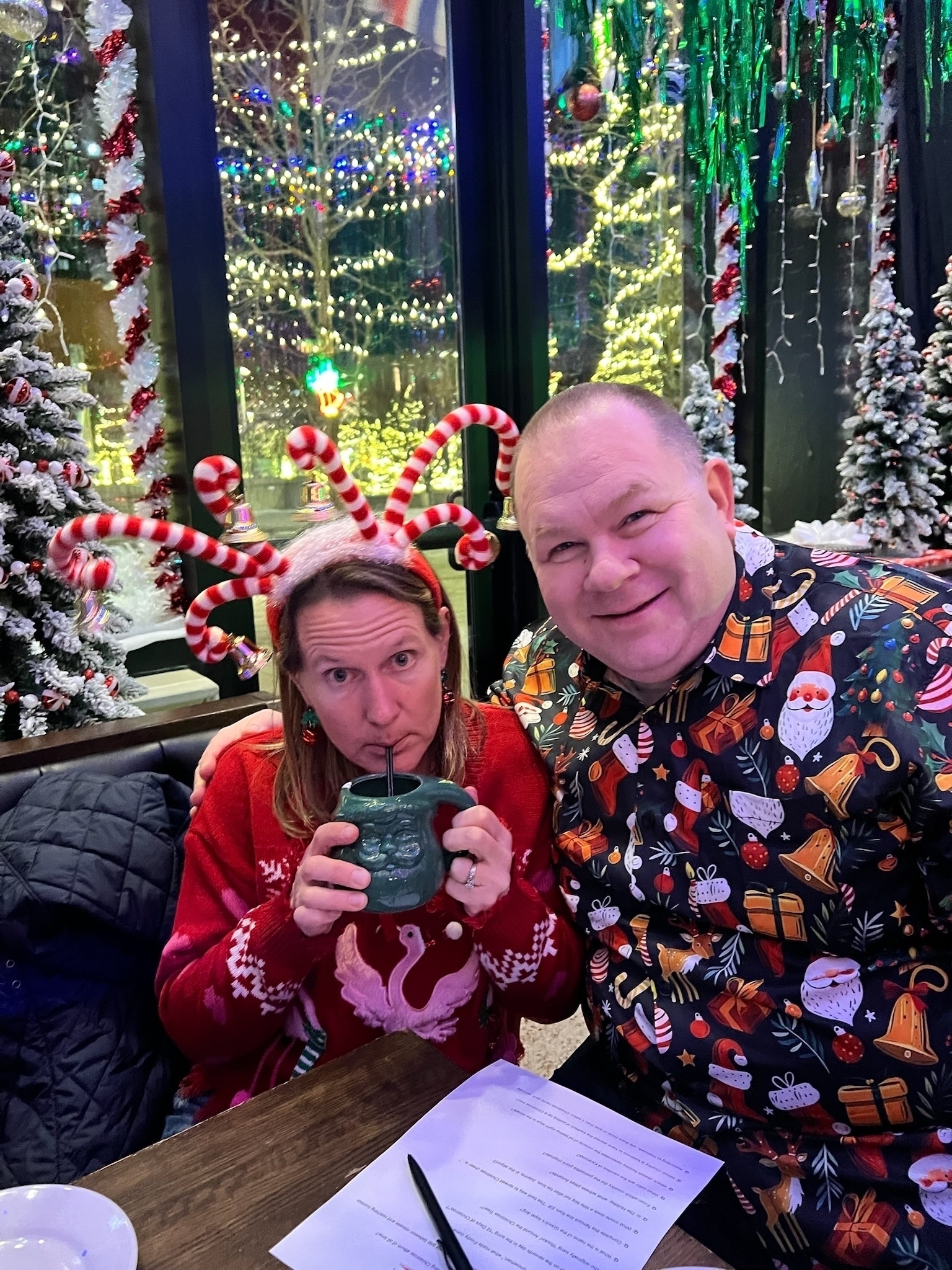 Auto-generated description: Two people in festive holiday attire, surrounded by decorated Christmas trees, are sitting at a table enjoying beverages.