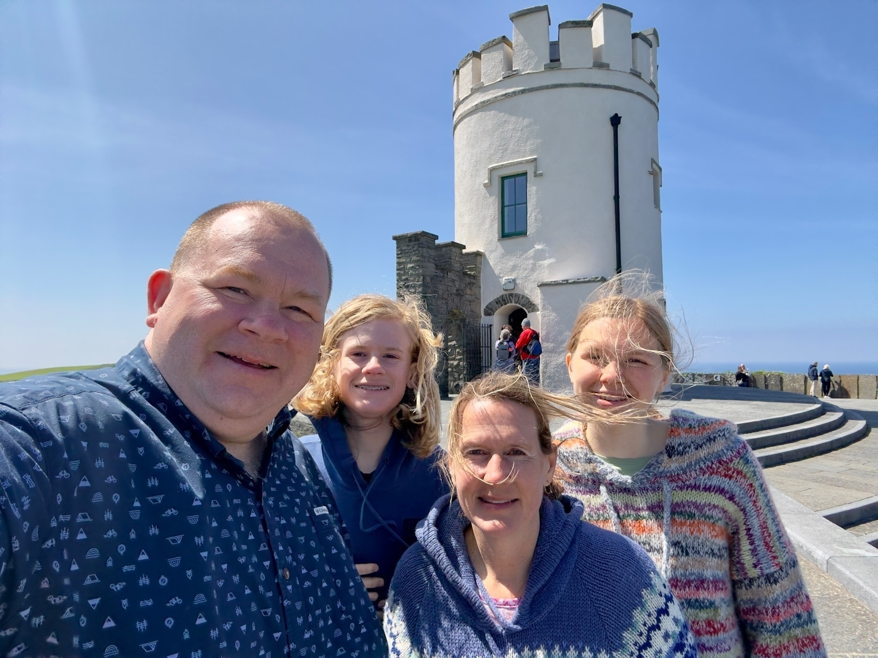 Auto-generated description: A family of four stands smiling in front of a circular stone tower on a sunny day.