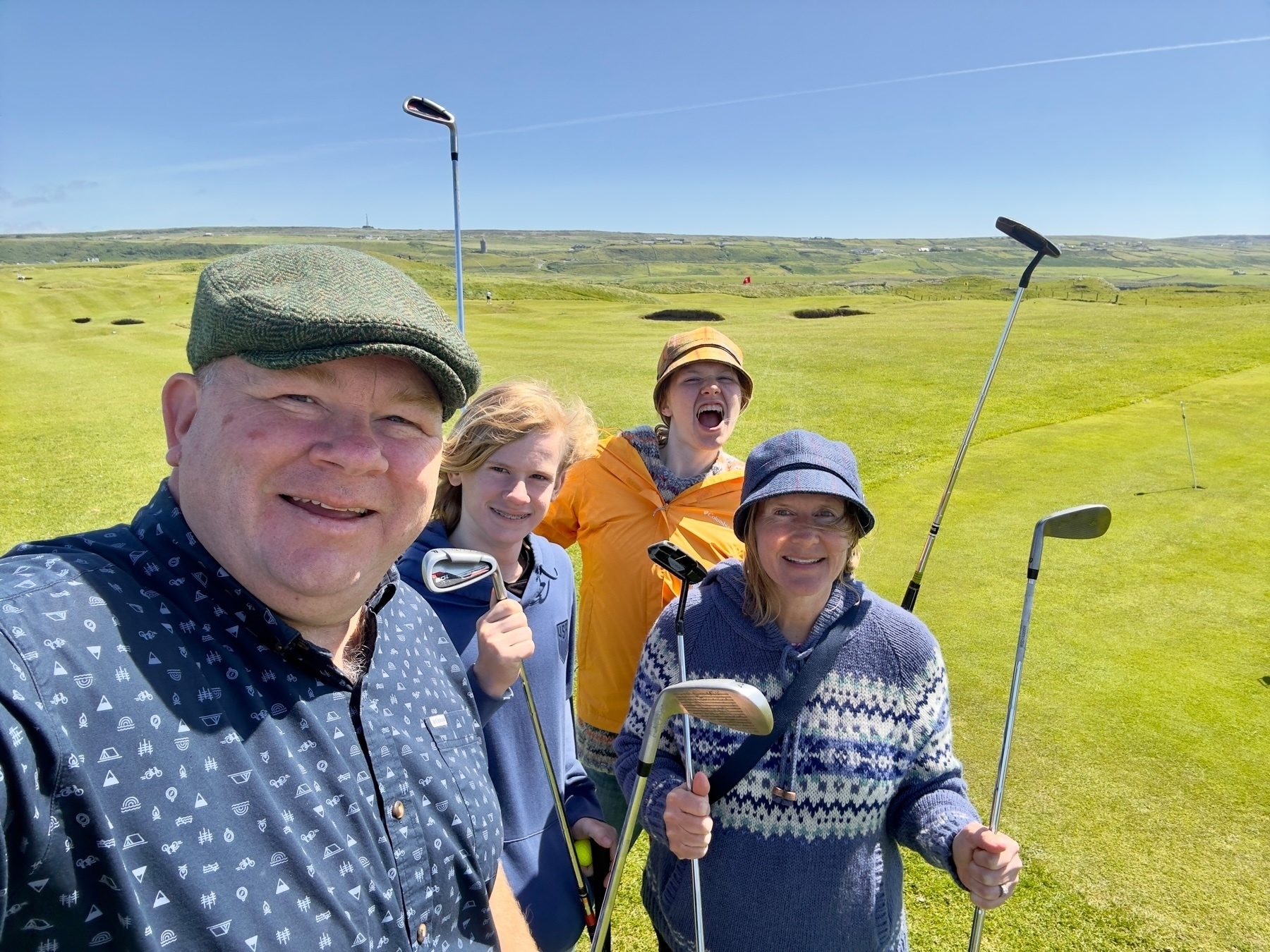 Auto-generated description: A smiling group of four people, each holding golf clubs, is standing together on a sunny golf course.