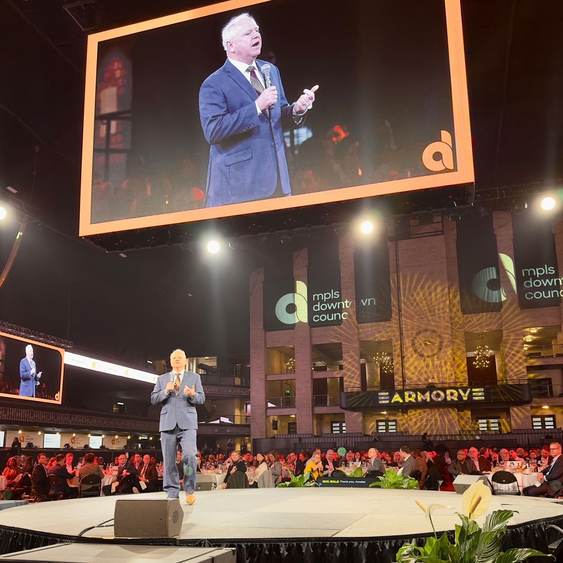 Governor Tim Walz speaking on stage at an event with an audience in the foreground. His image is also displayed on a large screen above him. The stage is adorned with floral arrangements, and banners with “mpls downtown council” logos are visible in the background