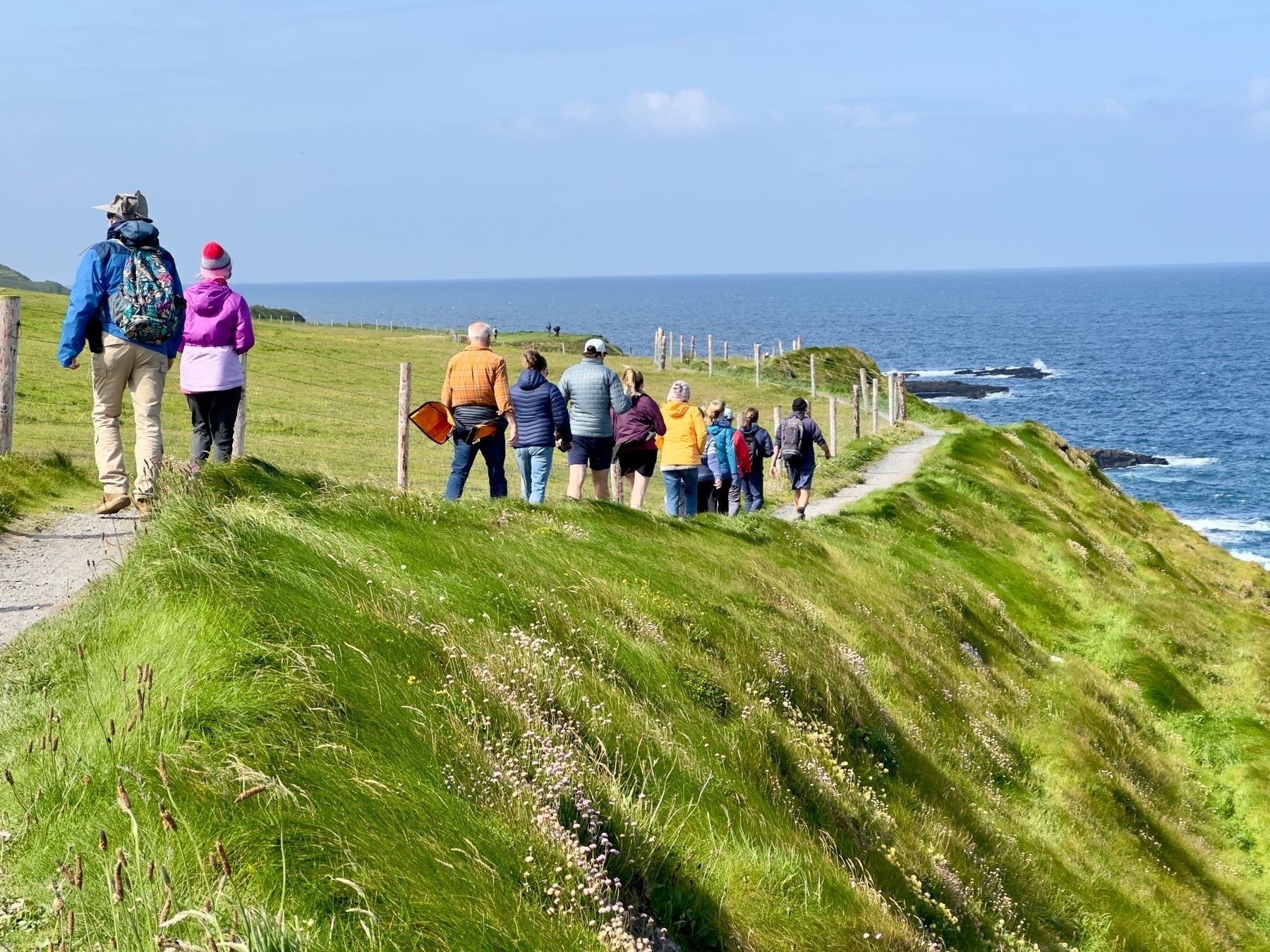 Auto-generated description: A group of people is walking along a grassy coastal path with the sea and blue sky in the background.