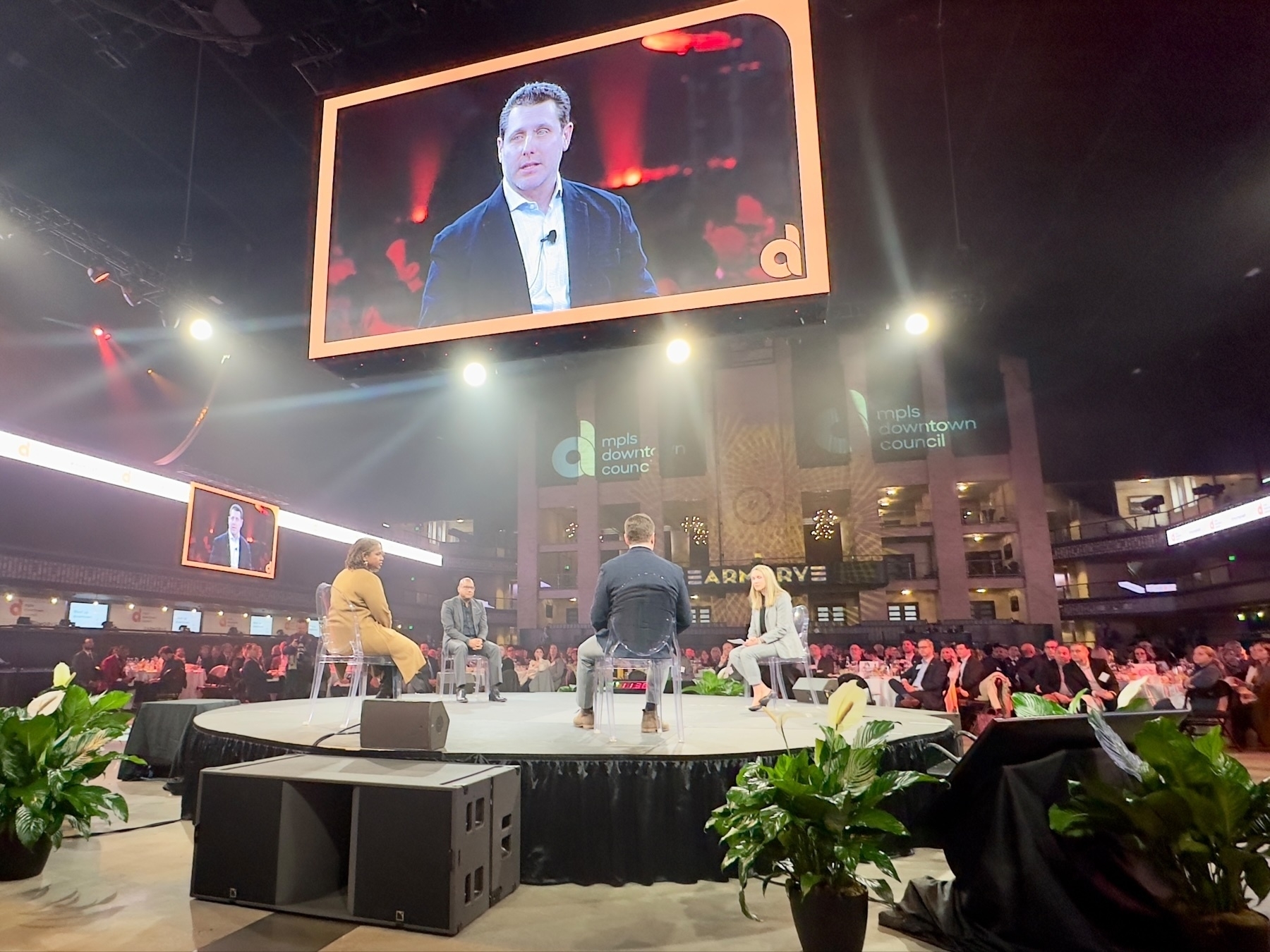 An event setting with a large audience and a stage where four people are seated, engaging in a discussion, with a large screen displaying a close-up of a speaker. The Minneapolis Downtown Council branding is visible in the background.