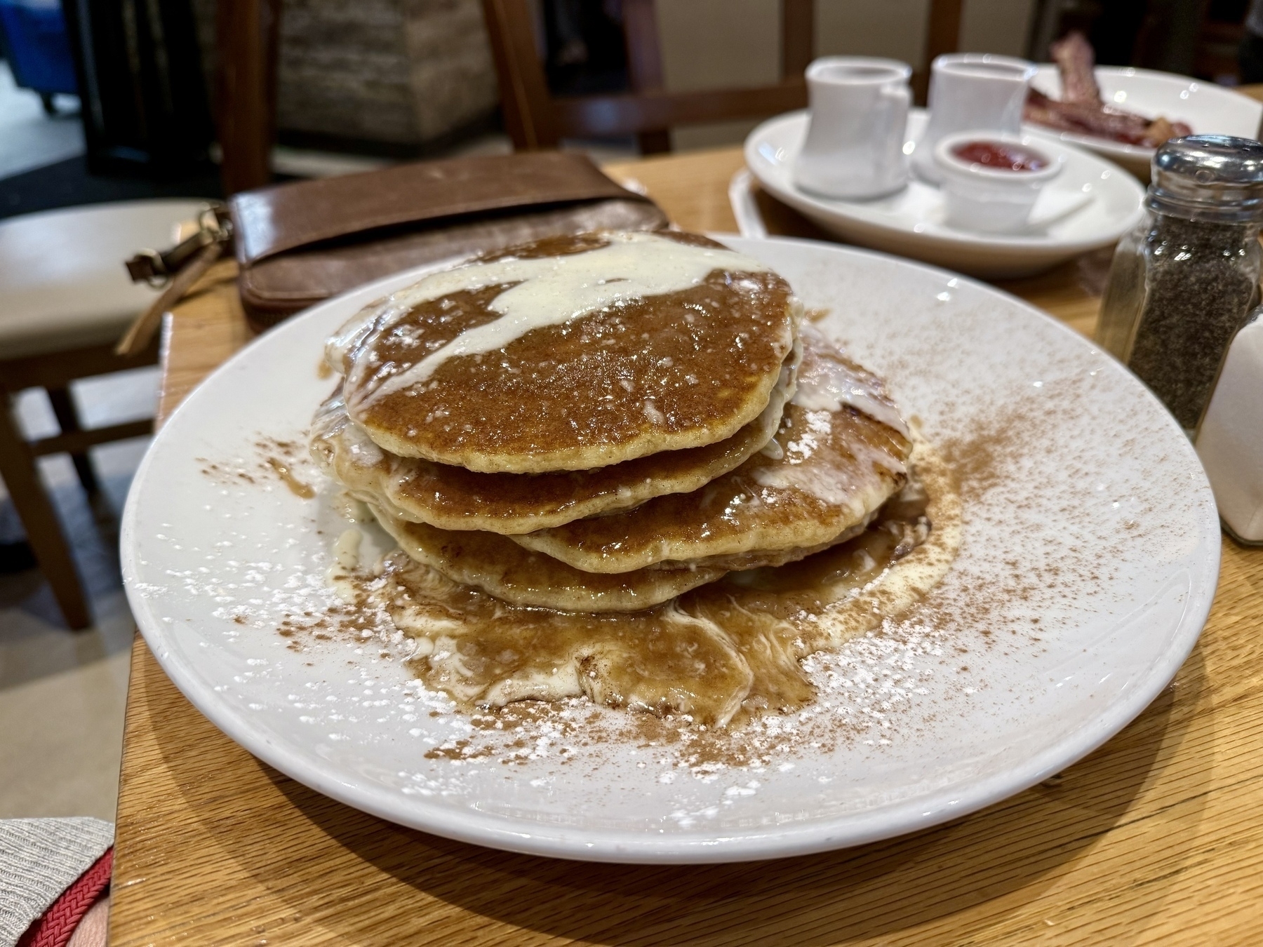 Auto-generated description: A plate of pancakes topped with syrup and powdered sugar sits on a wooden table, with two cups of sauce in the background.