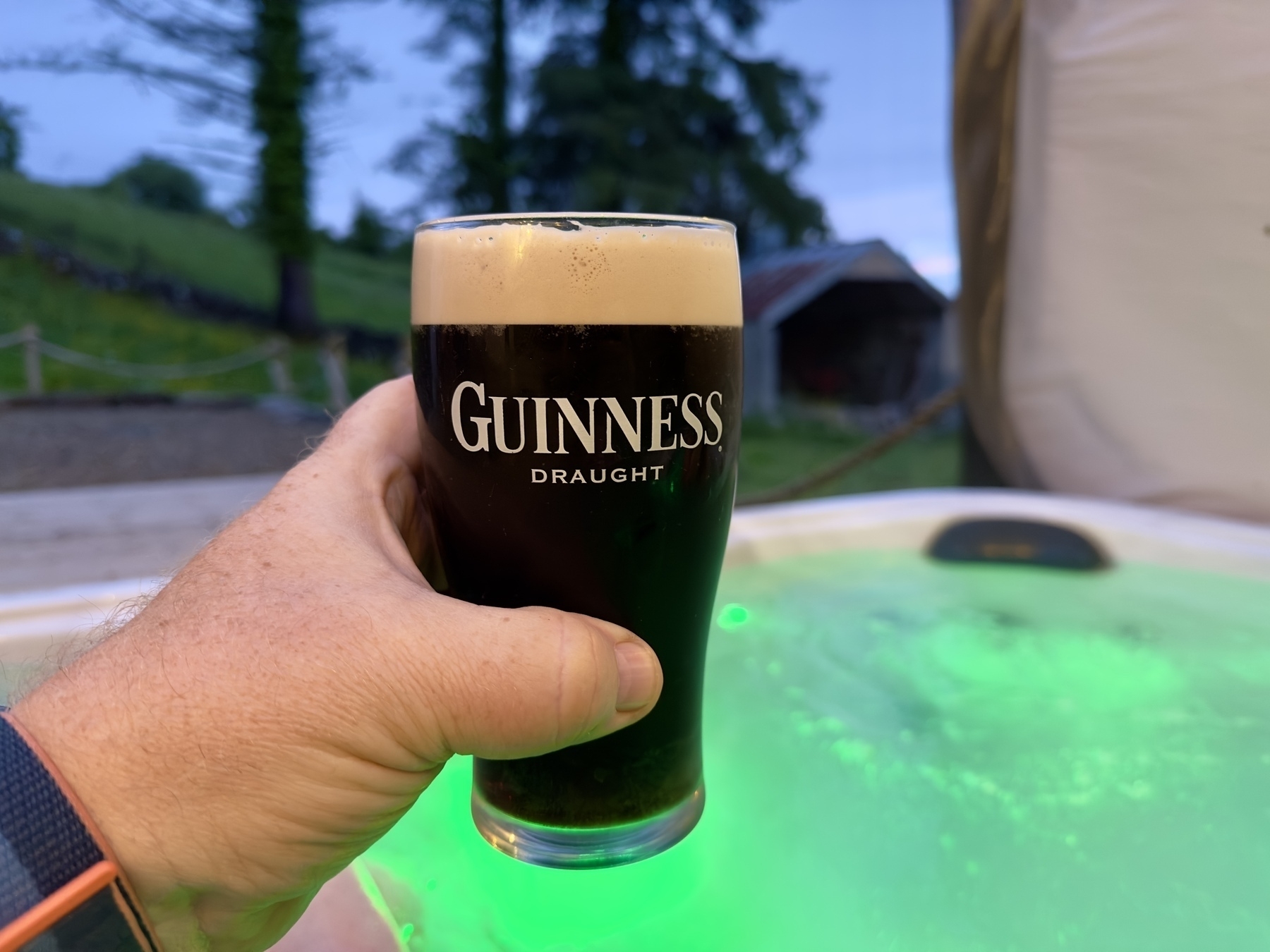 Auto-generated description: A hand is holding a pint of Guinness Draught near a glowing green hot tub in an outdoor setting.