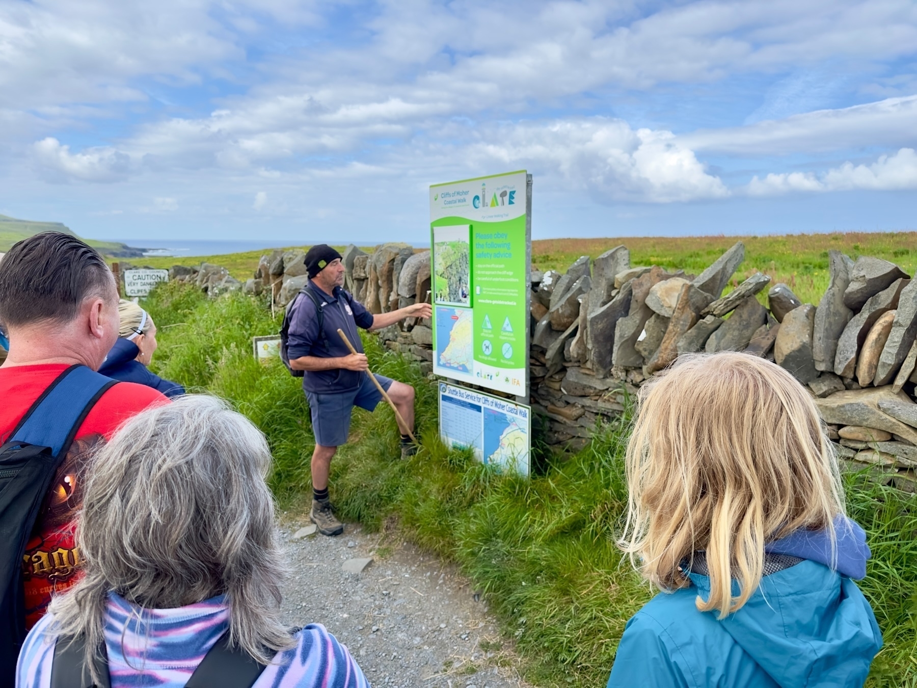 Auto-generated description: A group of people is gathered around a man giving a presentation in front of an informational sign outdoors, with a stone wall and grassy field in the background.