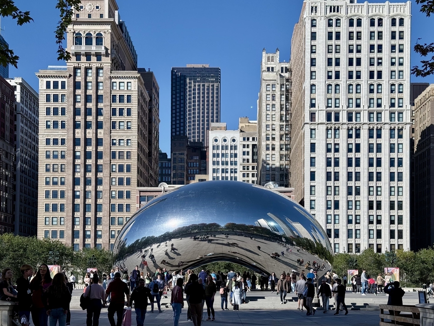 Auto-generated description: A large, reflective bean-shaped sculpture is surrounded by people and set against a backdrop of tall buildings in an urban park.