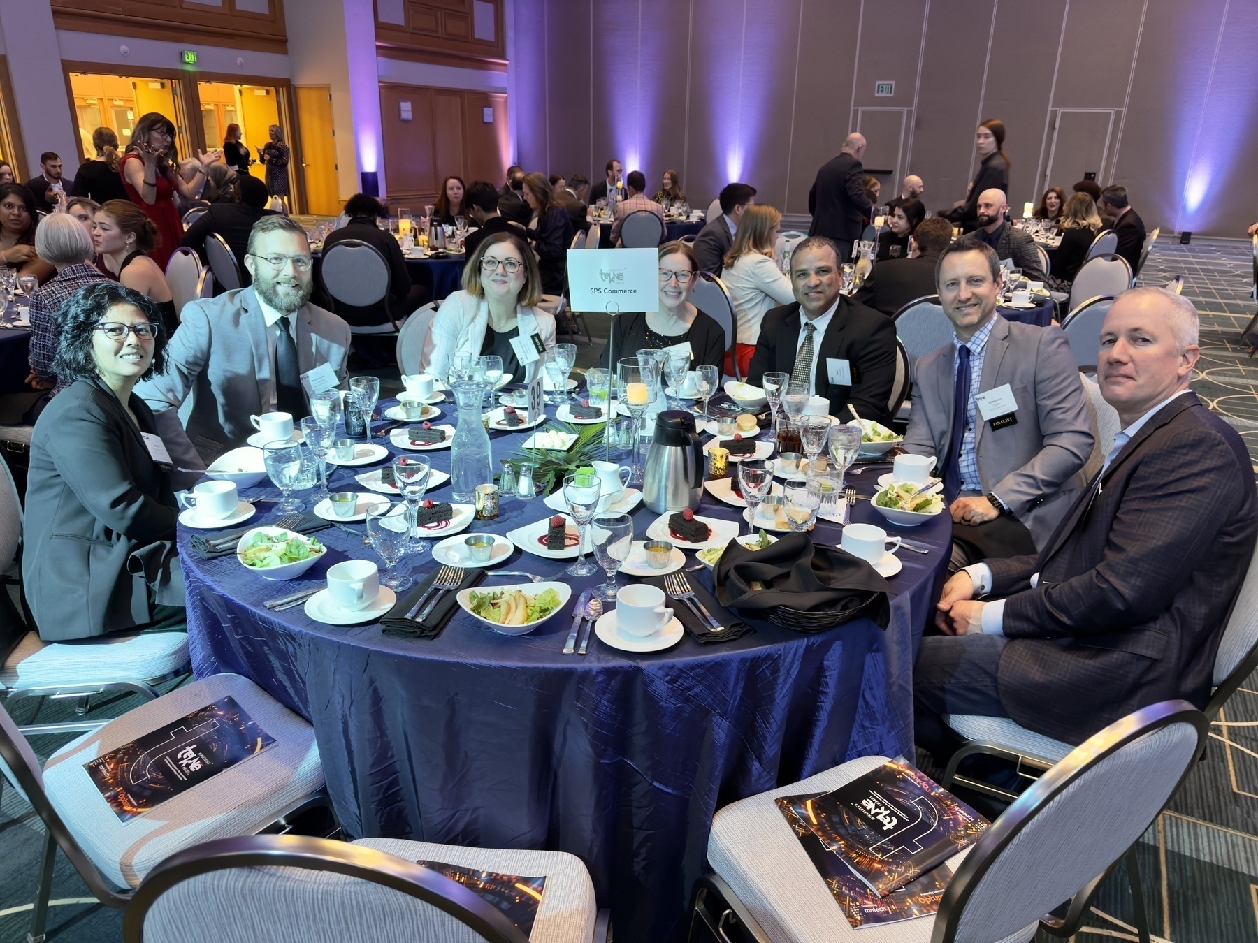 Auto-generated description: A group of people in formal attire are seated around a table set with dinnerware at an event.