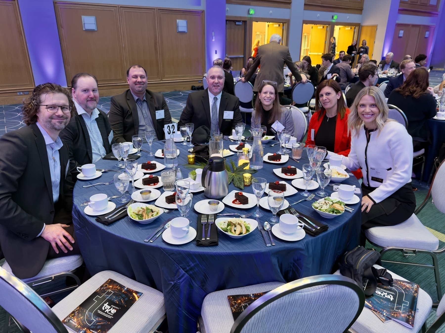 Auto-generated description: A group of people is seated around a table set for a formal banquet with salads, desserts, and beverages.