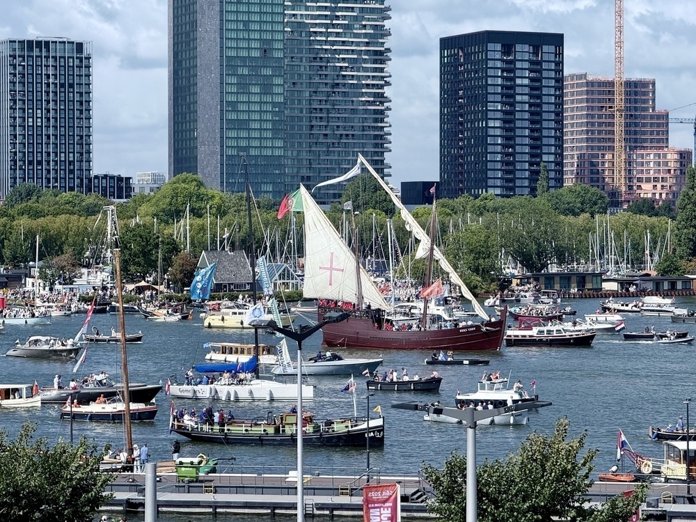 A bustling harbor scene with numerous boats, including a large sailing ship with a cross on its sail, set against a backdrop of modern city buildings and lush greenery.