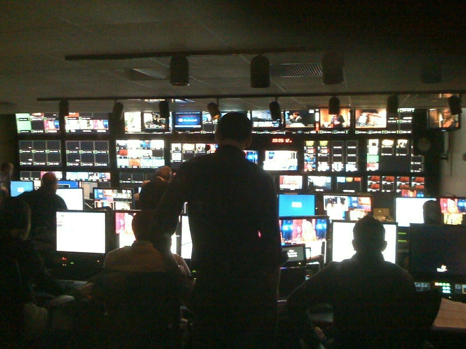 Auto-generated description: A busy television control room filled with multiple monitors and people working at computer stations.