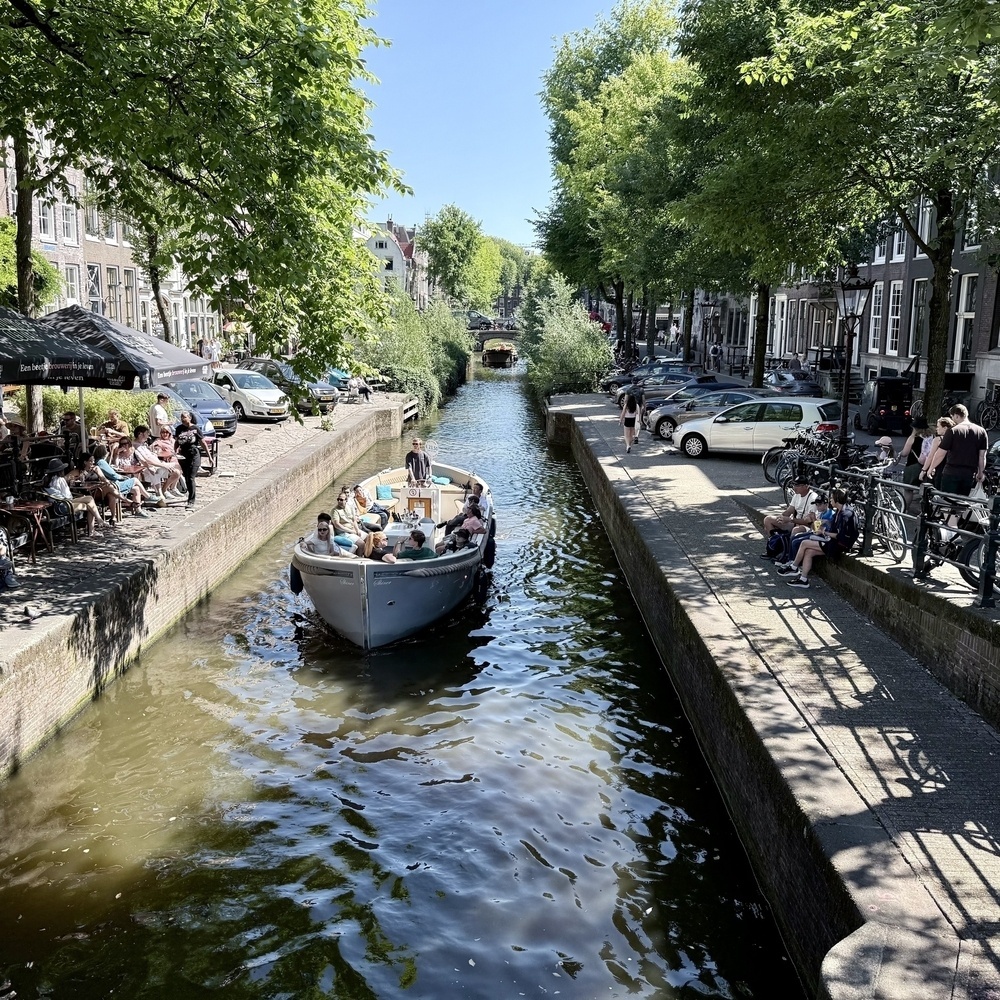 A boat filled with people travels along a tree-lined canal, with people sitting at a café and bicycles parked nearby.