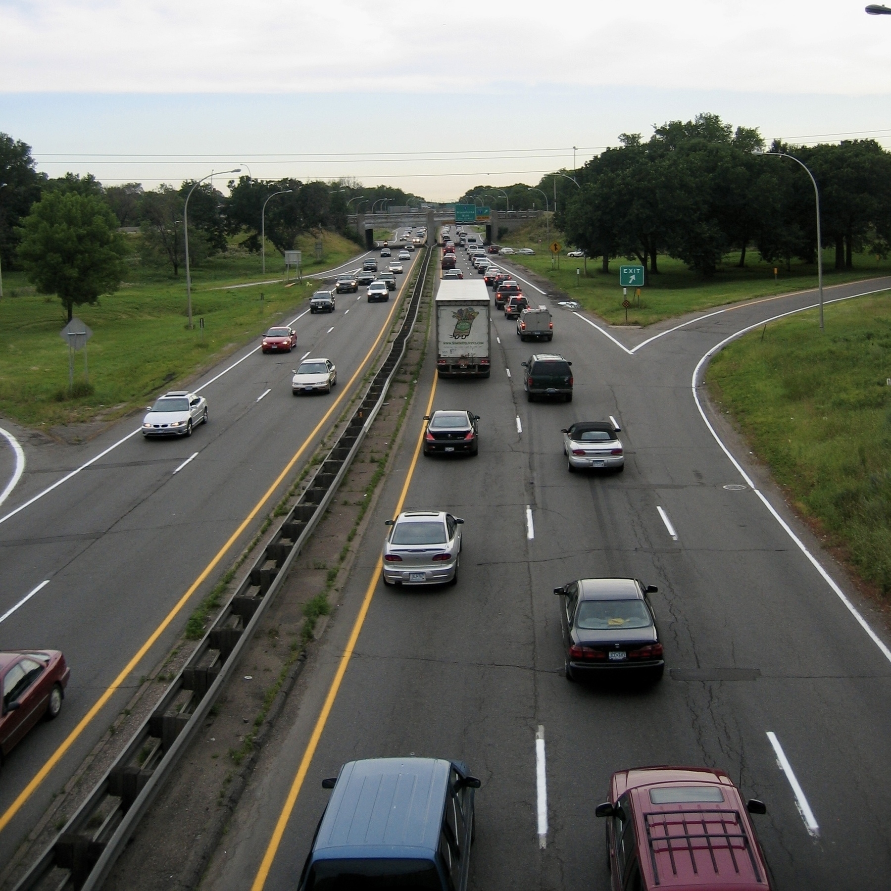 Auto-generated description: A busy highway features multiple lanes of traffic, including cars and a large truck, surrounded by green areas and trees.