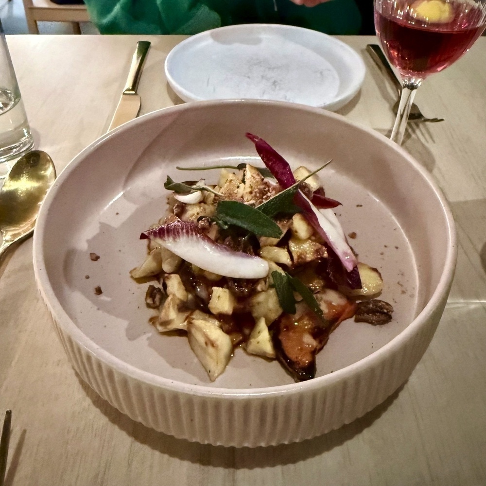 A plate of roasted vegetables and herbs is served next to a glass of red wine at a dining table.