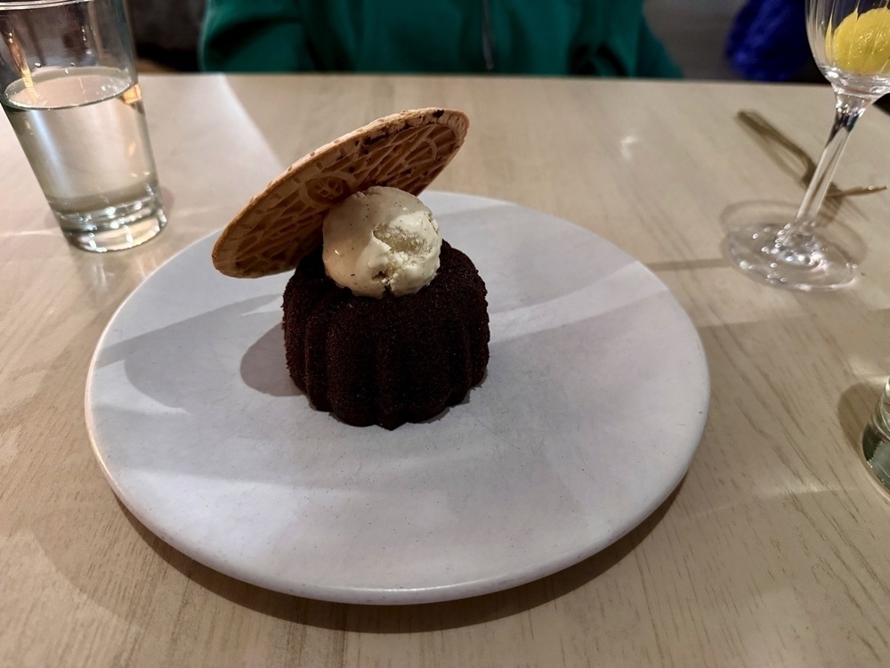 A chocolate dessert topped with a scoop of vanilla ice cream and a thin wafer sits on a white plate next to a glass of water and a wine glass.