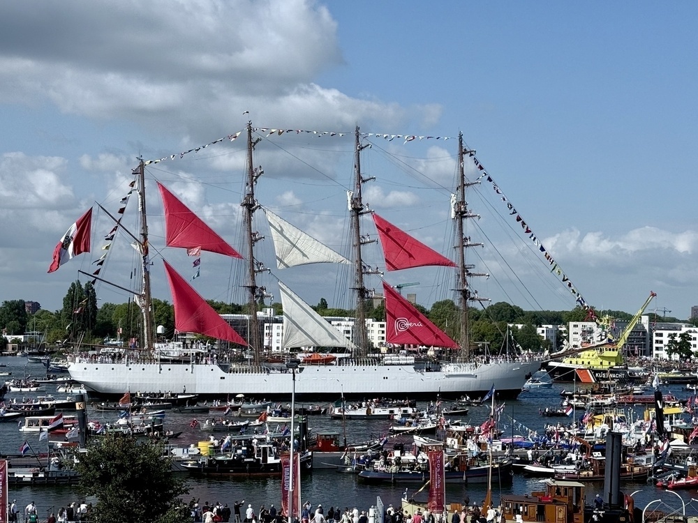 A tall ship with vibrant red sails is navigating a busy waterway surrounded by numerous smaller vessels and a crowd along the shore.