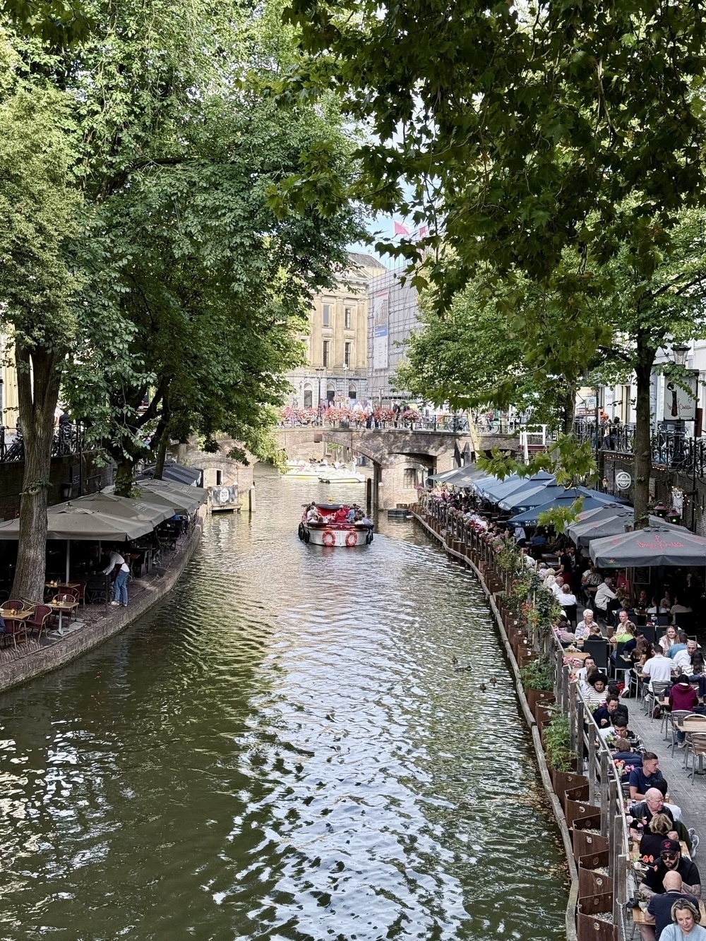 A canal lined with outdoor cafes is bustling with people and features a boat passing under a bridge.