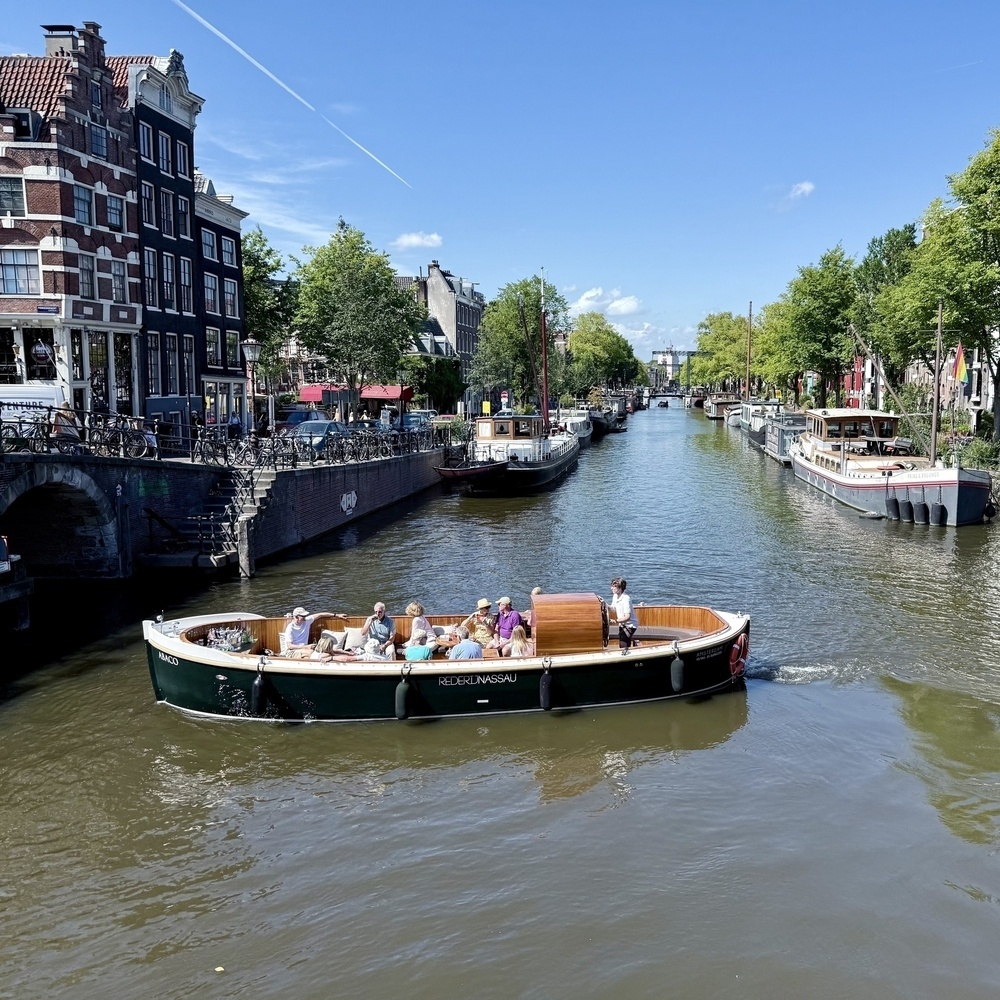 A boat cruises along a lively canal lined with historic buildings and lush trees under a clear blue sky.