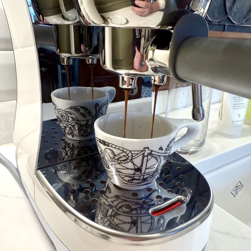 A coffee machine is brewing espresso into two decorative cups on a reflective surface.