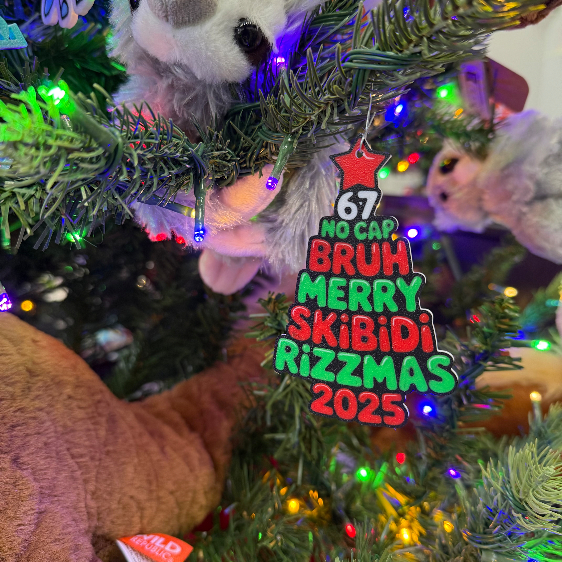 A Christmas tree is decorated with a humorous ornament featuring the text 67 No Cap Bruh Skibidi Rizzmas 2023 in colorful letters.