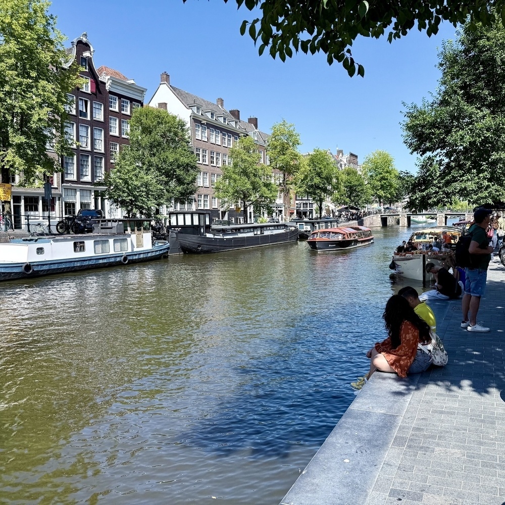A serene canal in a city is lined with boats and historic buildings, with people relaxing by the water's edge under a clear blue sky.