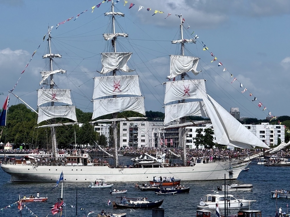 A large sailing ship with multiple masts and white sails is surrounded by smaller boats on a crowded waterway.