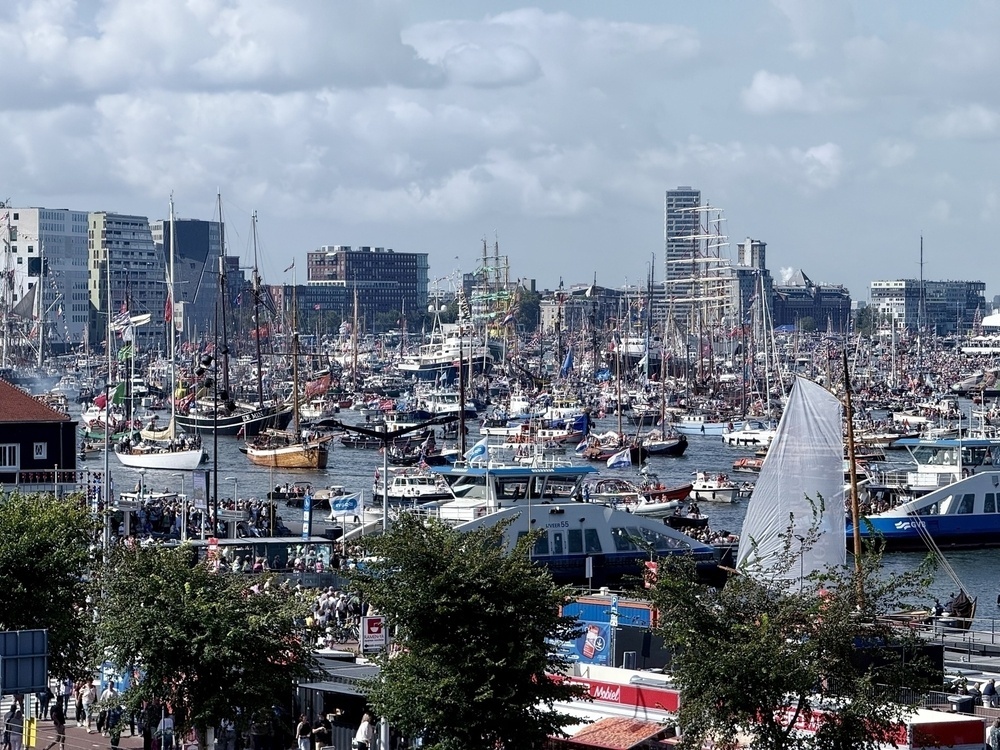 A bustling harbor is filled with numerous sailboats and ships surrounded by buildings under a partly cloudy sky.