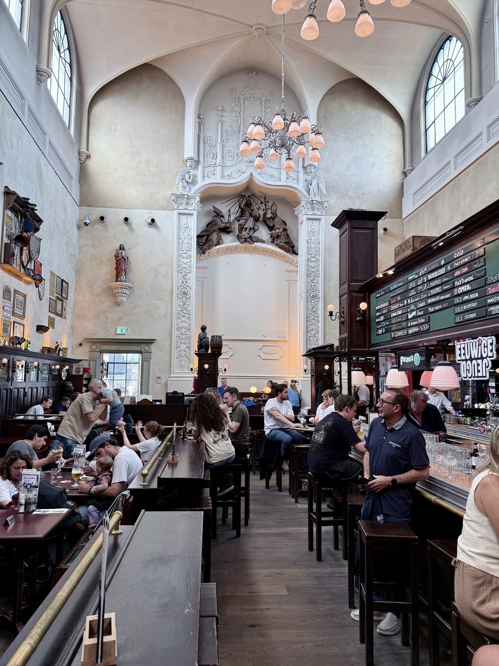 A spacious and ornate pub interior features high ceilings, decorative wall carvings, and patrons dining and socializing at tables and the bar.
