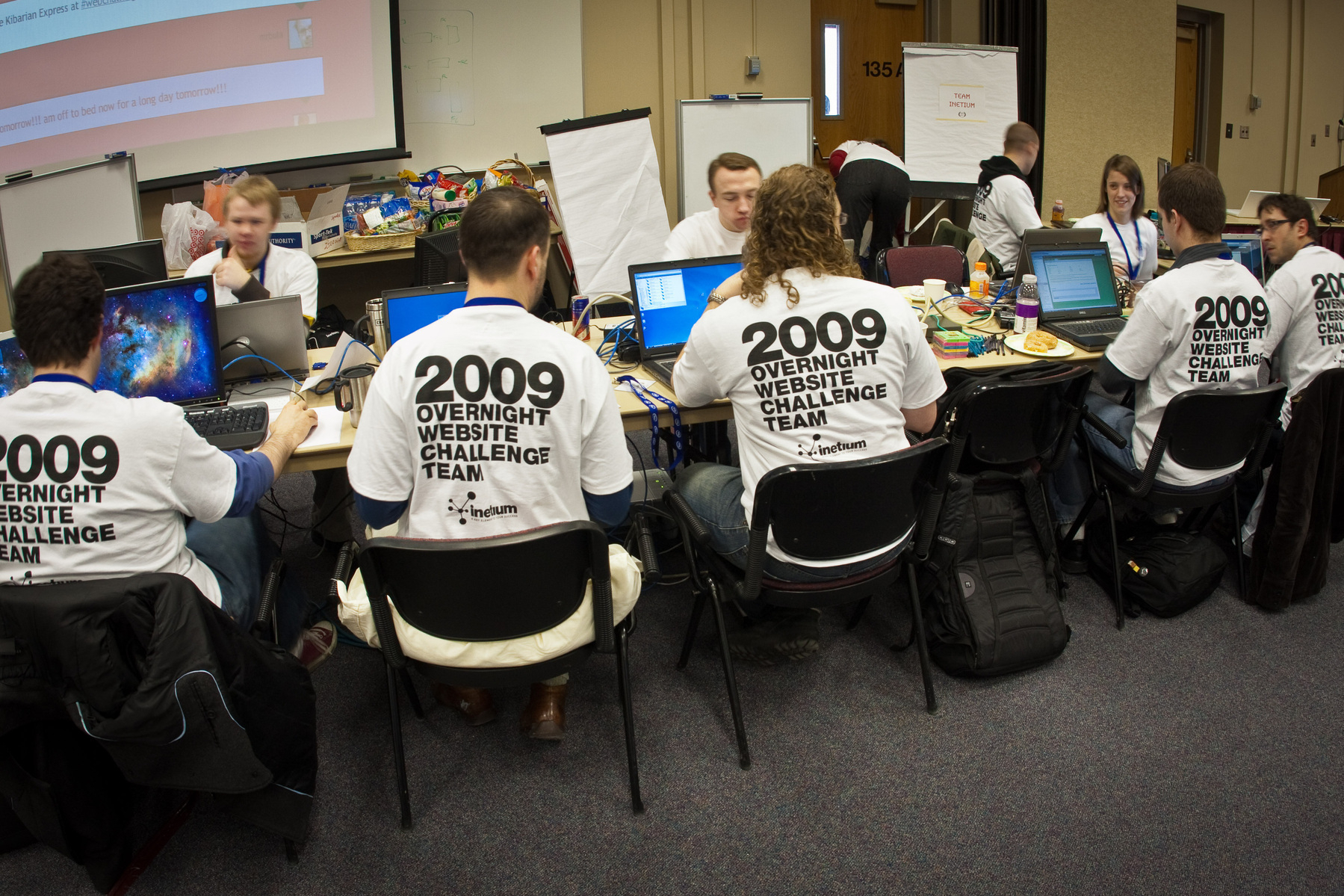 Auto-generated description: A group of people wearing matching 2009 Overnight Website Challenge Team shirts are seated around a table with laptops and computers, engaged in a collaborative work session.