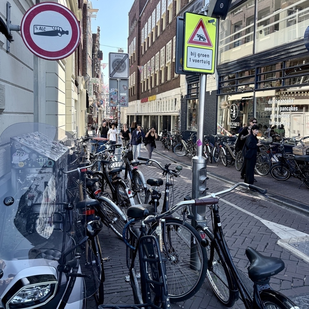 A narrow, bustling street features rows of bicycles parked alongside pedestrians walking past shops and signs, creating a typical urban scene.