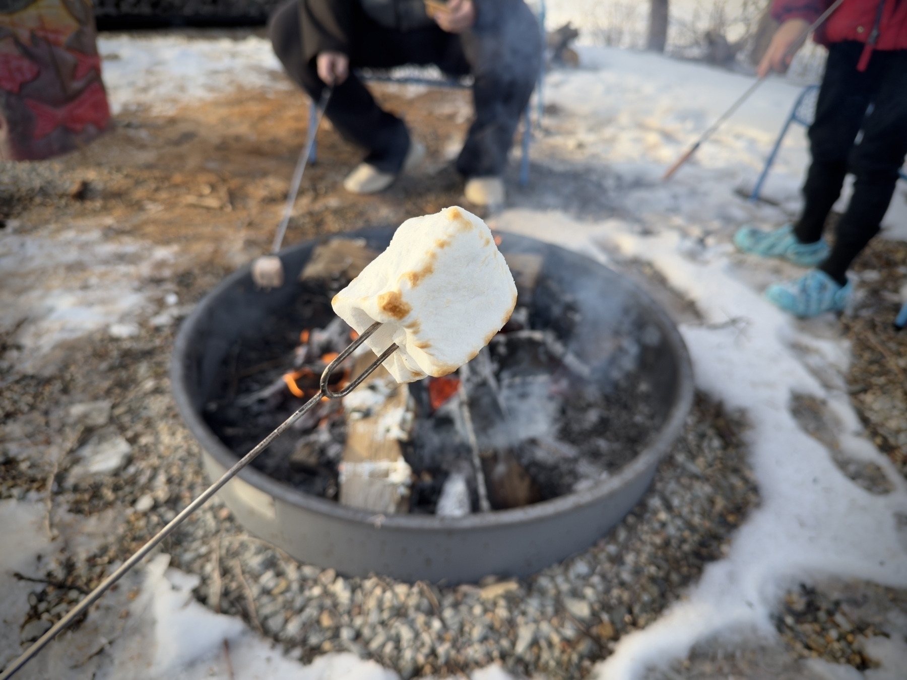 A marshmallow is being roasted on a stick over an outdoor fire pit while two people are seated nearby.