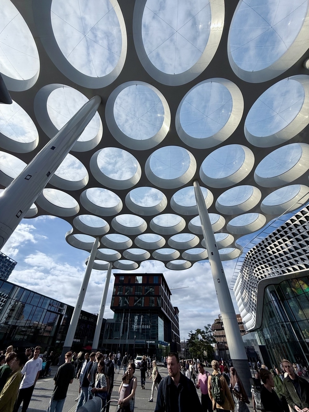 People walk under a large, modern canopy structure with circular cutouts in an urban setting.