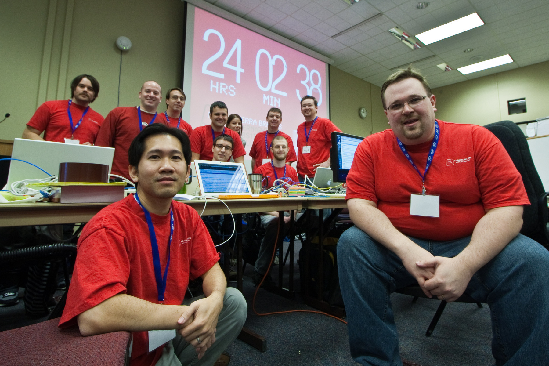 Auto-generated description: A group of people wearing red shirts with name tags are posing in front of a digital timer displaying 24 HRS 02 MIN.