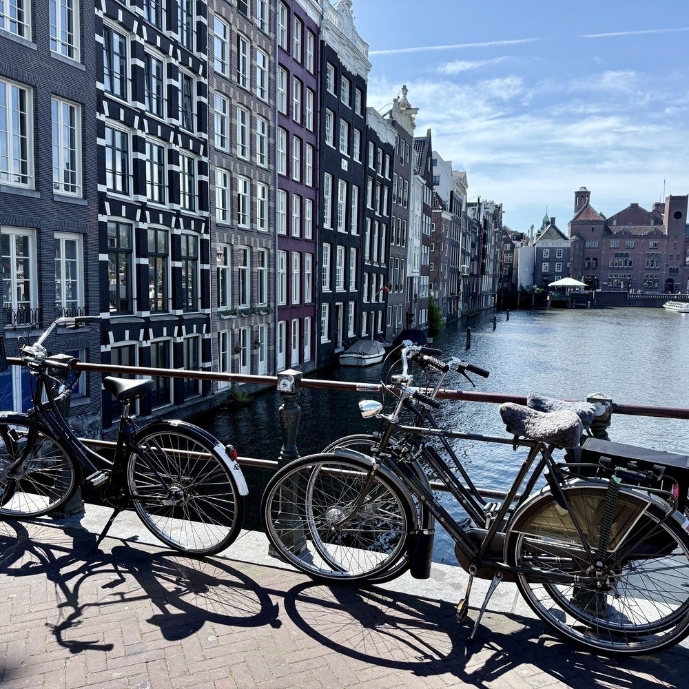 Bicycles are parked along a canal in front of traditional, narrow Dutch buildings under a clear blue sky.