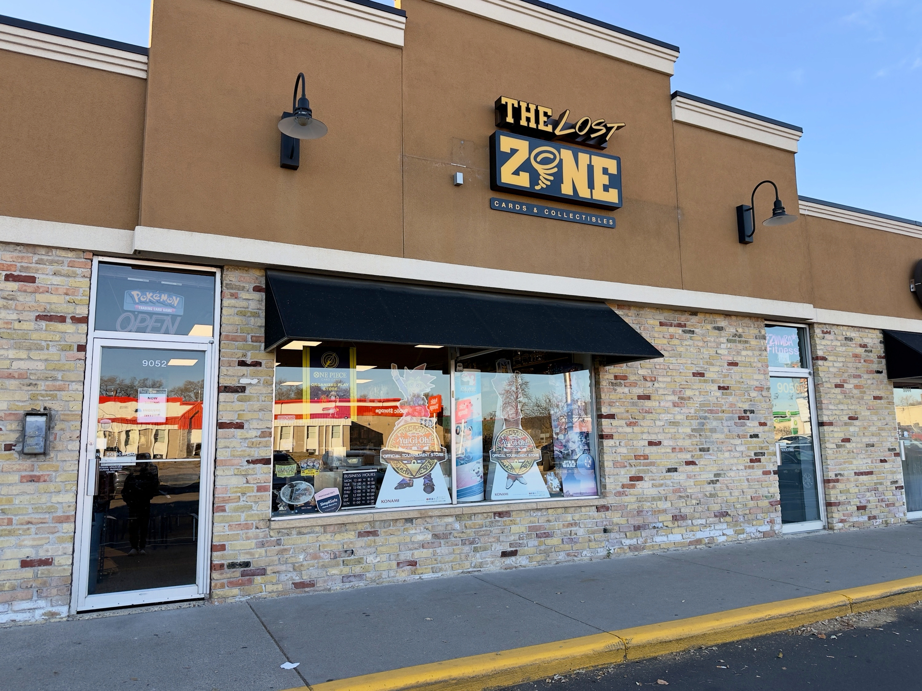 A storefront called The Lost Zone features brickwork, a black awning, and signage indicating it's a games and collectibles shop.