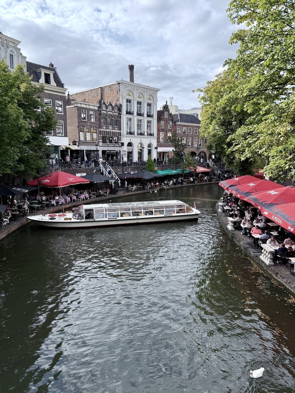 A canal lined with outdoor cafes and historic buildings features a boat passing by under a cloudy sky.