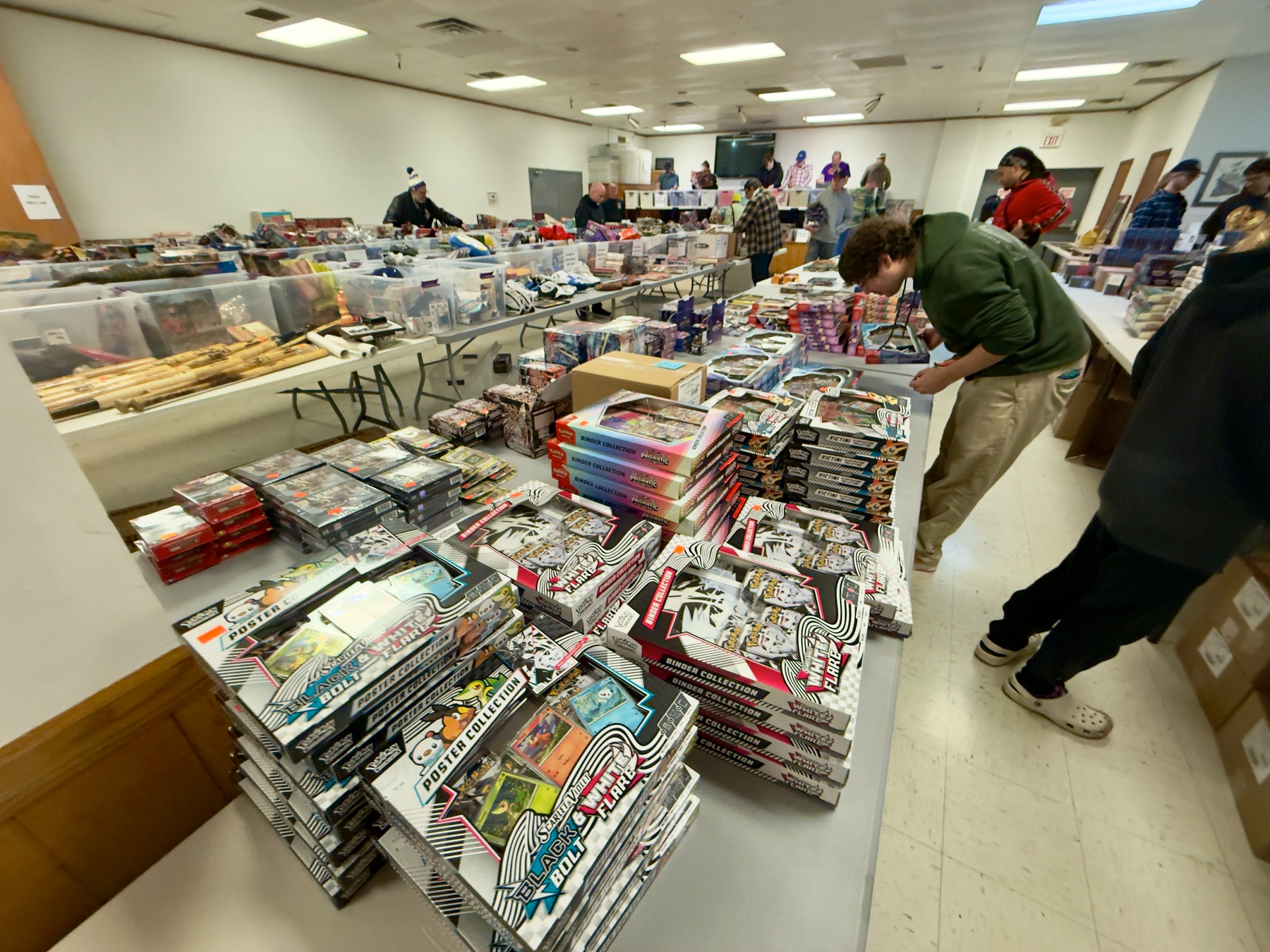 A large room is filled with tables displaying numerous collectible trading card packages, with several people browsing through them.