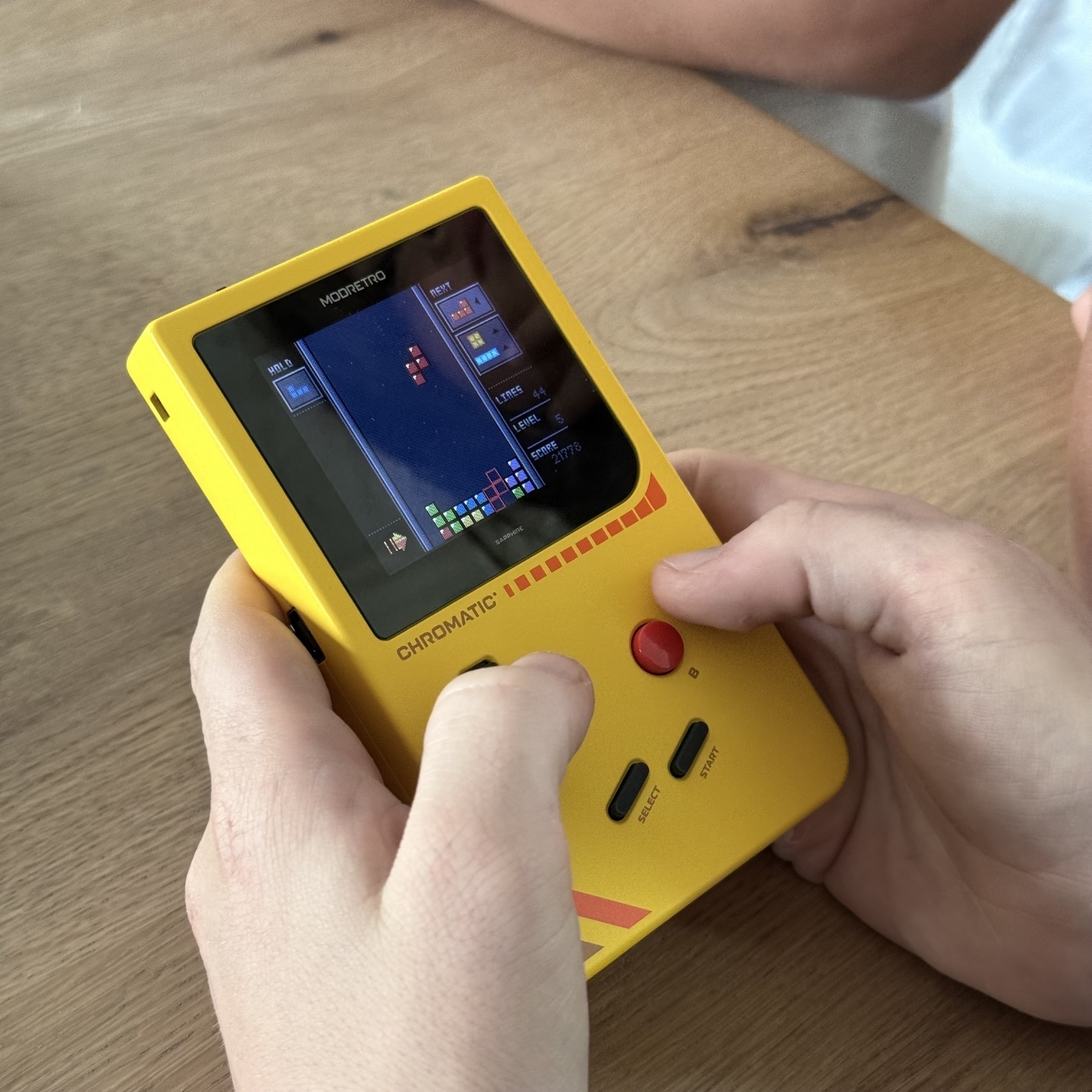 A pair of hands is holding a yellow handheld gaming device displaying a Tetris game.