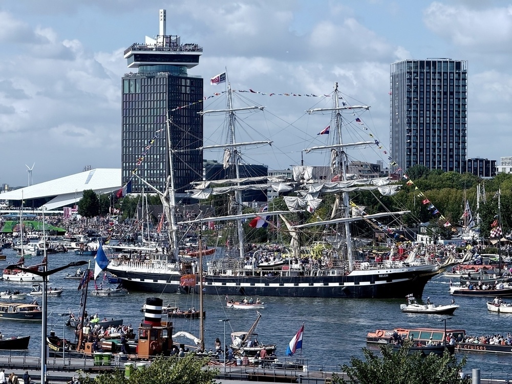 A large sailing ship adorned with flags is surrounded by numerous smaller boats on a bustling waterway, set against an urban backdrop with tall buildings and a cloudy sky.