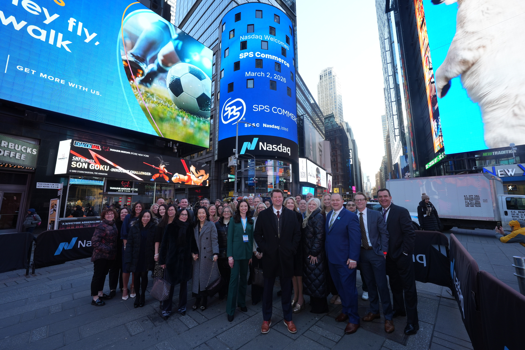 A group of people is gathered in Times Square in front of large digital billboards, including a prominent Nasdaq display.